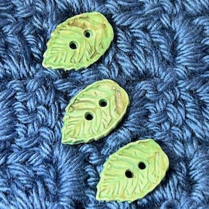 May include: Three green leaf-shaped buttons with two holes each. The buttons have a textured surface and are arranged on a blue knitted fabric.