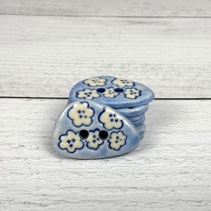 May include: A stack of light blue ceramic buttons with a rounded triangular shape. Each button features a repeating pattern of small, off-white five-petaled flowers with dark blue centers and two holes for sewing in the center of the design.