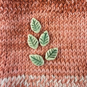 May include: Five small, pale green ceramic leaf-shaped buttons with dark green painted vein details. Each button features a pointed tip and a rounded base, arranged in a cluster against a textured, variegated orange and pink knitted fabric background.