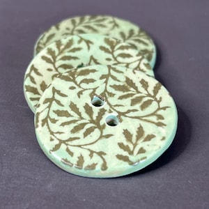 May include: Three round, ceramic buttons with a light green background and a brown leaf pattern. Each button has two holes for sewing. The buttons are stacked slightly, showing the design and texture.