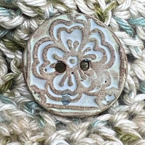 May include: A light blue ceramic button with a raised floral design. The button has two holes and a brown glaze.