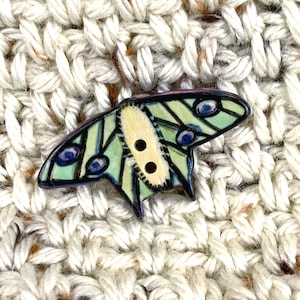 May include: A decorative butterfly button with a light green and blue wing design. The button has two holes and a cream-colored center. The button is set against a textured, off-white fabric background.