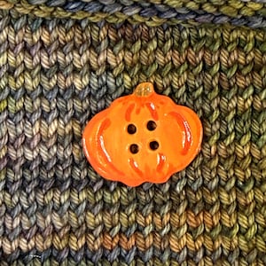May include: An orange pumpkin-shaped button with four black button holes. The button is glossy and sits on a background of knitted fabric in shades of green, brown, and blue. A seasonal craft supply.