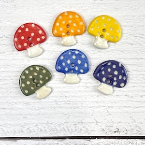 May include: A set of six mushroom-shaped buttons in red, orange, yellow, green, light blue, and dark blue. Each button features white polka dots on the cap, a white stem, and two central holes for attachment, arranged in a scattered pattern.