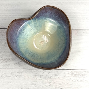 May include: A heart-shaped ceramic bowl featuring a reactive glaze with a deep purple and brown rim that transitions into a light cream and pale blue center. The interior surface displays a fine, radiating brushstroke pattern throughout the glaze.