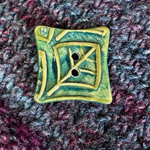 May include: Square ceramic button with a green and yellow design. The button has two holes and a leaf-like pattern in the center. The button is on a textured, multi-colored fabric background.