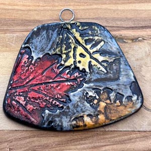 May include: A ceramic ornament with a black, gray, red, yellow, and orange leaf design. The ornament has a silver loop for hanging.