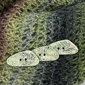 May include: Three light green ceramic buttons with black leaf designs and two holes each. The buttons are oval-shaped and rest on a textured, multi-colored knitted fabric in shades of green, purple, and gray.