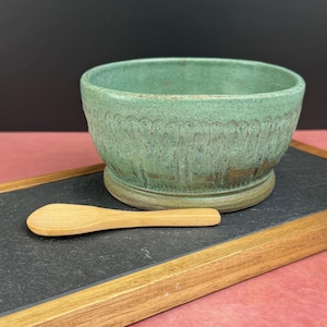 May include: A green ceramic bowl with a decorative rim and a wooden spoon rest on a black surface. The bowl has a textured finish and a pedestal base. The wooden spoon is light-colored and rests on a black slate-like surface.