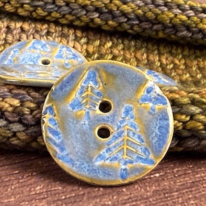 May include: Close-up of ceramic buttons with a blue and gold glaze. Each button features a raised design of a pine tree. The buttons have two holes for sewing and are set against a textured background.