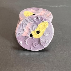 May include: Stack of ceramic buttons with a floral design. The buttons are round, with a speckled purple base, and feature yellow and pink accents. Each button has two holes for sewing.