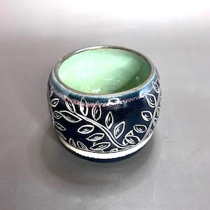 May include: A handcrafted ceramic bowl with a dark blue exterior and a light green interior. The bowl features a white leaf design around the exterior. The rim has a silver-colored trim. The bowl is approximately 3 inches in diameter and 2.5 inches tall.