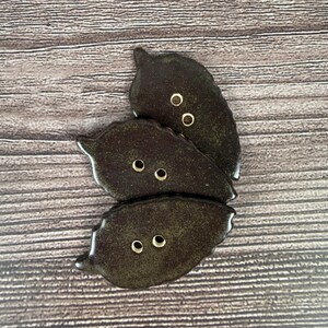 May include: Three dark brown leaf-shaped buttons with gold-colored eyelets are arranged on a wooden surface. The buttons have a glossy finish and a textured appearance, suggesting a handcrafted or artisan design. The buttons are likely for sewing or crafting projects.
