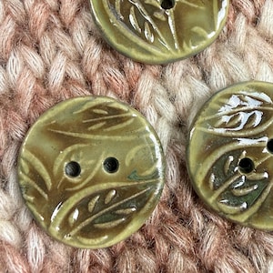 May include: Three round, olive green ceramic buttons featuring a glossy finish and a raised, embossed leaf pattern. Each button has two central holes for attachment, with subtle variations in the glaze color and texture across the surface.