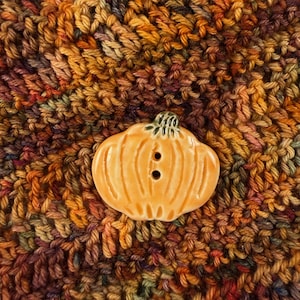 May include: A ceramic button shaped like a pumpkin with a glossy orange finish and a dark green stem. The button features two vertical holes in the center for attachment and has a smooth, rounded surface with carved ridges representing the pumpkin.