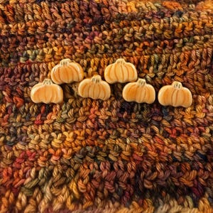 May include: Six small, light orange ceramic pumpkin-shaped buttons with vertical ridges. Each button features a smooth, glossy finish and a rounded, segmented design, arranged in two rows against a textured, multi-colored yarn background.