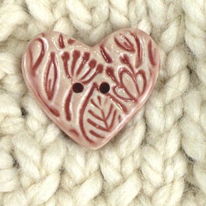 May include: A heart-shaped button with a floral design in shades of pink and red. The button has two holes and is set against a background of thick, off-white knitted fabric. The button is approximately 1 inch in width.
