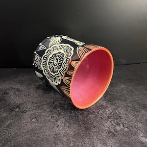 May include: A cylindrical ceramic cup featuring a dark navy blue exterior with a white etched floral pattern. The interior of the cup is a vibrant, solid coral pink color. The rim is finished with a thin, matching coral pink band around the edge.