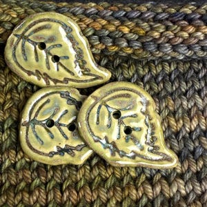 May include: Close-up of three leaf-shaped ceramic buttons in shades of yellow-green with brown and blue accents. The buttons have two holes for sewing and are set against a textured, multi-colored knitted background.