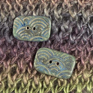 May include: Two rectangular ceramic buttons featuring a glossy finish with a blue and green wave pattern. Each button has two central holes for attachment, showcasing a smooth, glazed surface with intricate, curved line details throughout.