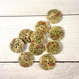 May include: A collection of ten round ceramic buttons featuring embossed leaf patterns in shades of sage green, tan, and burnt orange. Each button has two central holes and a textured surface with detailed botanical motifs of various foliage.
