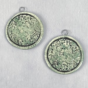 May include: Two round, ceramic pendants with a light green background and black tree designs. Each pendant has a silver-colored loop at the top for hanging. The pendants are approximately 1.5 inches in diameter and feature a textured border.