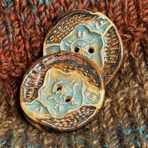 May include: Two ceramic buttons with a teal and brown floral design. The buttons have two holes for sewing and are set against a textured, multicolored fabric background. The buttons are round and have a glossy finish.