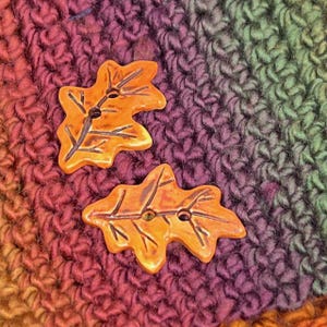 May include: Two orange ceramic buttons shaped like oak leaves. The buttons have a textured surface and are on a multi-colored crocheted background.