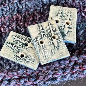 May include: Three rectangular, light gray ceramic buttons with a textured, architectural design. Each button has two holes for sewing. The buttons rest on a background of dark blue, purple, and red knitted fabric.