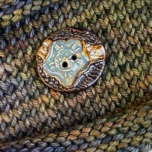 May include: A close-up of a ceramic button with a floral design and two holes. The button is light blue and brown, with a textured surface. The button is attached to a knitted fabric in shades of green, brown, and gray.