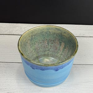 May include: A ceramic bowl featuring a smooth, light blue exterior and a speckled interior glaze that transitions from a muted green to a warm, earthy brown tone. The rim is defined by a dark, metallic-toned edge, creating a distinct contrast.