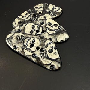 May include: Three elongated, oval-shaped buttons featuring a distressed black and off-white finish. Each button displays a repeating pattern of detailed skull motifs with hollow eye sockets and teeth, integrated with two central sewing holes.