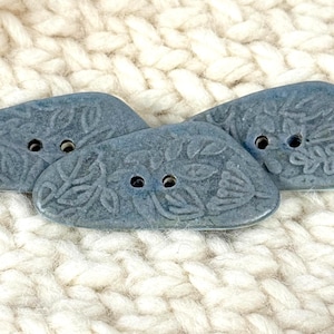 May include: Close-up of three blue ceramic buttons with floral designs. Each button has two holes for sewing. The buttons are set against a textured, off-white background, suggesting a craft or sewing project.