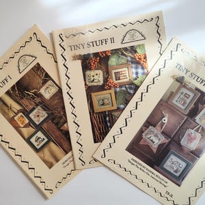 May include: Three cream-colored paper booklets titled Tiny Stuff I, Tiny Stuff II, and Tiny Stuff, each featuring a decorative black border and photographic illustrations of small framed cross-stitch projects, berries, and wooden textures.