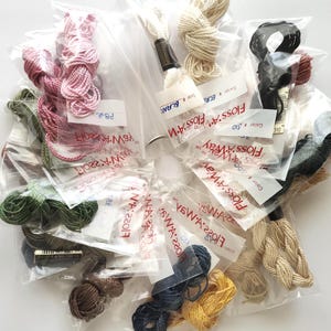 May include: A collection of embroidery floss in various colors, including pink, green, black, and yellow, packaged in clear plastic bags. Each bundle is labeled with color information and brand details. The floss is tightly wound and ready for use.