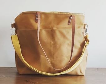 May include: A tan canvas tote bag with a yellow shoulder strap and brown leather handles. The bag has a rectangular shape with two exterior pockets and a zipper closure. The bag is sitting on a wooden surface.