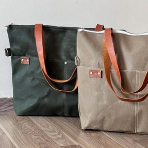 K&ouml;nnte beinhalten: Zwei Canvas-Tragetaschen, eine dunkelgr&uuml;ne, die andere beige. Beide haben braune Ledergriffe und eine Vordertasche. Die Taschen haben einen Rei&szlig;verschluss und ein kleines Lederetikett mit einem Logo.