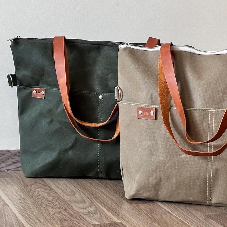 K&ouml;nnte beinhalten: Zwei Canvas-Tragetaschen, eine dunkelgr&uuml;ne, die andere beige. Beide haben braune Ledergriffe und eine Vordertasche. Die Taschen haben einen Rei&szlig;verschluss und ein kleines Lederetikett mit einem Logo.