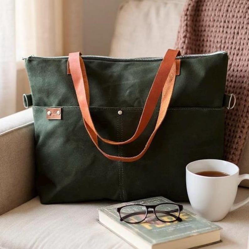 K&ouml;nnte beinhalten: Eine olivgr&uuml;ne Canvas-Tragetasche mit braunen Ledergriffen und Akzenten. Die Tasche hat einen Rei&szlig;verschluss und eine Vordertasche. Eine wei&szlig;e Tasse Tee, ein Buch und eine Brille befinden sich im Vordergrund.