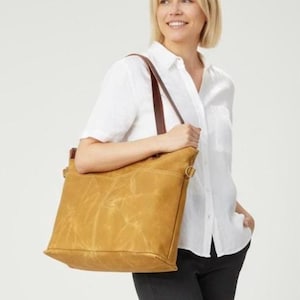 Könnte beinhalten: Eine senffarbene Tragetasche mit braunen Lederriemen. Die Tasche hat eine rechteckige Form mit strukturiertem Aussehen und einer Seitentasche. Die Tasche wird über der Schulter getragen.