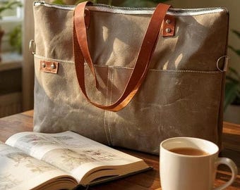 May include: A canvas tote bag in shades of brown and gray with brown leather handles. The bag is on a wooden table next to an open book and a white mug of tea on a wooden coaster. The bag has a zipper closure.