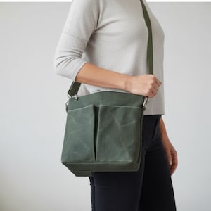 May include: Olive green messenger bag with two front pockets and a long adjustable strap. The bag is made of a waxed canvas material and has silver-toned hardware. The bag is being worn over the shoulder.