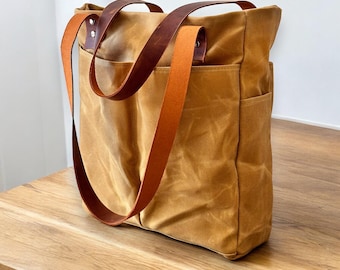 May include: A mustard-colored tote bag with brown leather handles and straps. The bag has two exterior pockets and a spacious interior. The leather straps are attached with rivets. The bag is made of a durable, waxed canvas material.