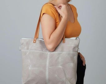 May include: A light gray tote bag with brown leather handles and a white zipper. The bag has two front pockets and a silver metal ring on the side. The bag is made of a durable, textured material.