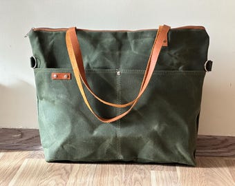 May include: A large, olive green tote bag with brown leather handles and a zipper closure. The bag has two exterior pockets and metal hardware. The bag is made of a durable canvas material, suitable for carrying various items.
