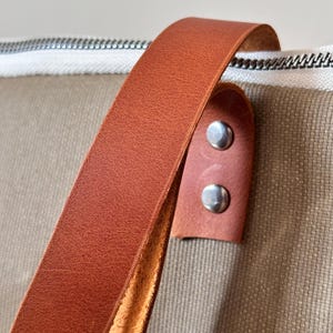 K&ouml;nnte beinhalten: Nahaufnahme einer beigefarbenen Canvas-Tasche mit einem braunen Ledergriff. Der Griff ist mit silbernen Nieten an der Tasche befestigt. Der Rei&szlig;verschluss ist wei&szlig; mit einem schwarzen Streifen.
