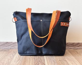 May include: A black canvas tote bag with brown leather handles and accents. The bag has a zippered top, exterior pockets, and a white stitched seam down the front. The bag is sitting on a wooden floor.