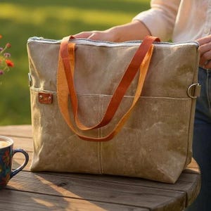 K&ouml;nnte beinhalten: Eine gro&szlig;e, braune Tragetasche mit orangefarbenen Ledergriffen. Die Tasche hat einen Rei&szlig;verschluss und ein kleines Lederetikett. Die Tasche steht auf einem Holztisch. Der Hintergrund ist verschwommen, mit einem Hauch von Gr&uuml;n.