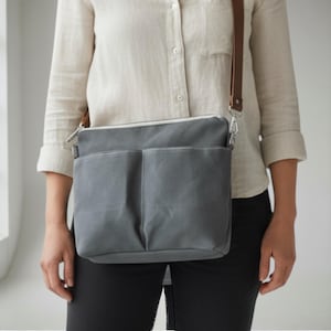 Könnte beinhalten: Graue Canvas-Umhängetasche mit zwei Vordertaschen und einem braunen Lederriemen. Die Tasche hat einen silbernen Reißverschluss und ein weißes Innenfutter. Die Tasche wird über der Schulter getragen.
