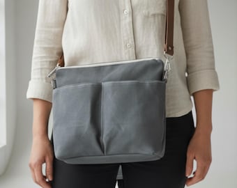May include: A gray canvas crossbody bag with two front pockets and a brown leather strap. The bag has a silver zipper and a white interior lining. The bag is worn over the shoulder.